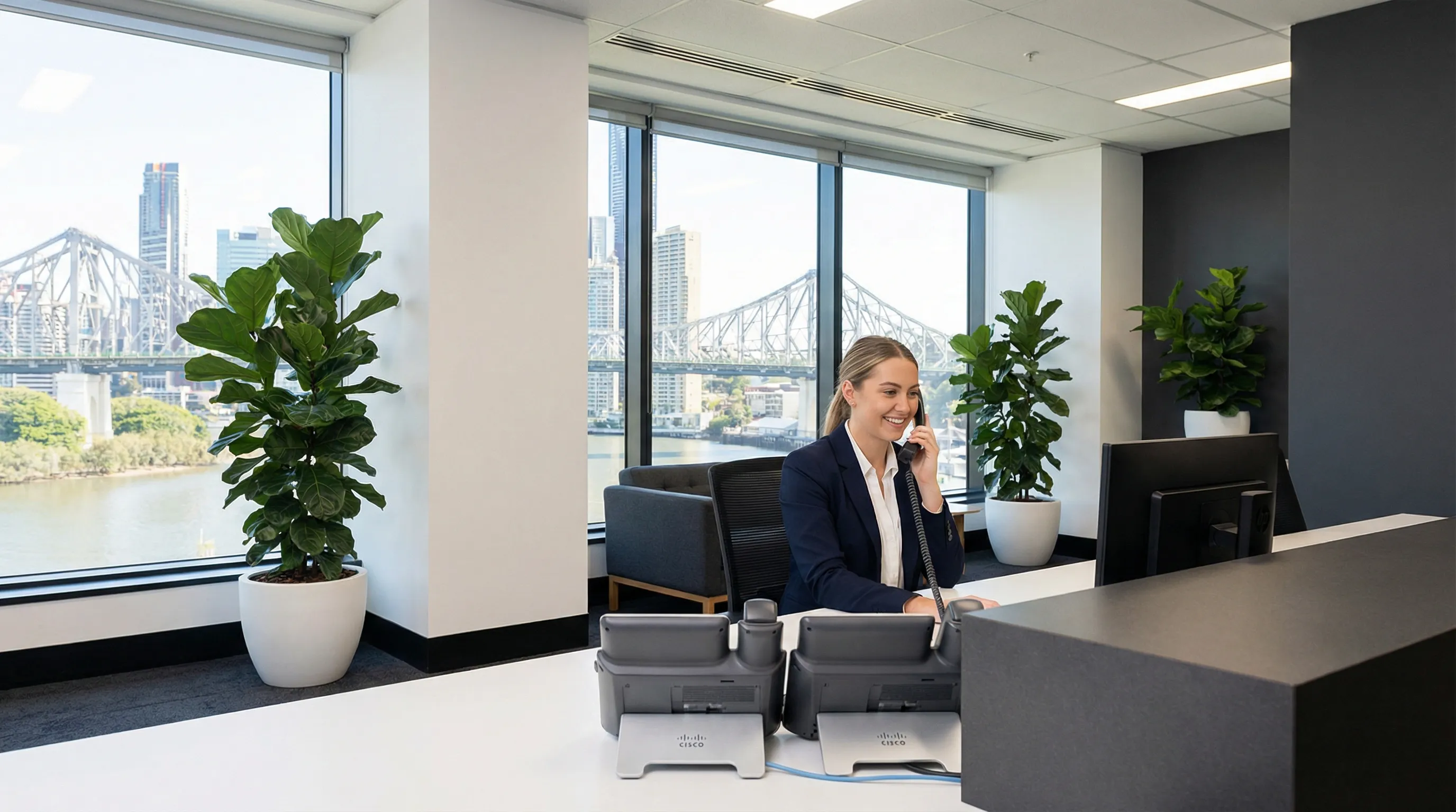 A friendly Australian receptionist answers a modern multi-line desk phone in a bright Brisbane office with city views